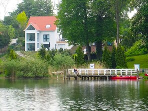 A house with a red roof, a dock extending into a body of water, and a person sitting on the dock. - am Seeufer by Interhome (Malente-Gremsmühlen)
