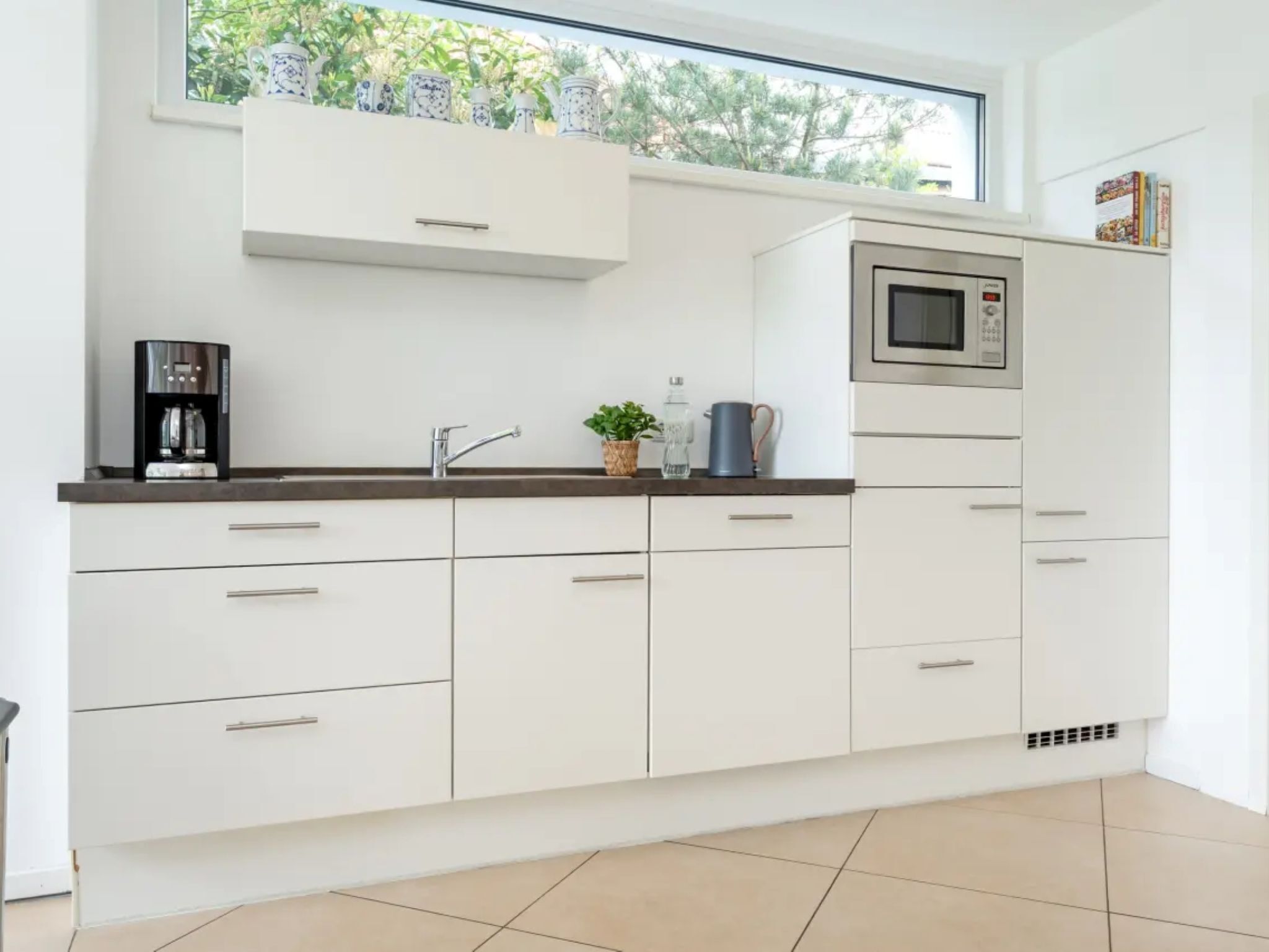 Een moderne keuken met witte kastjes, een koffiezetapparaat, een magnetron en een raam met uitzicht op groen.