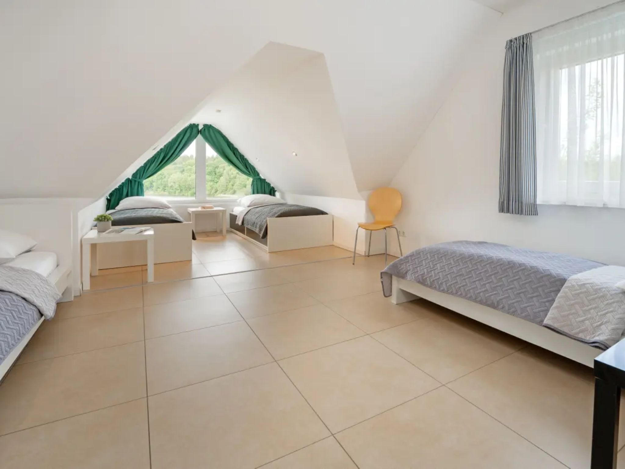 Une chambre avec un plafond en pente, deux lits, une chaise et une fenêtre avec des rideaux.