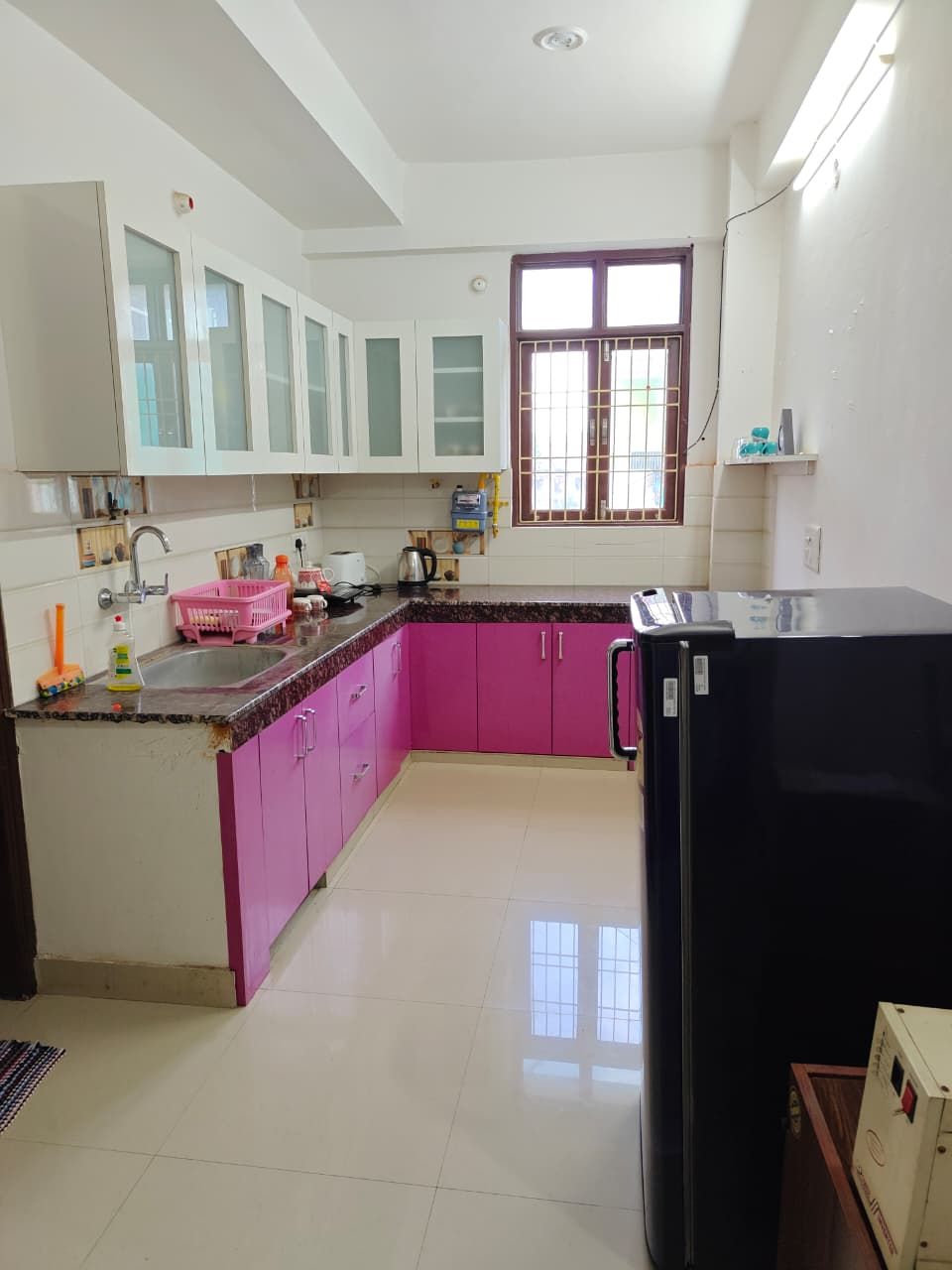 A kitchen with white cabinets, a pink countertop, a black refrigerator, and a window with bars.