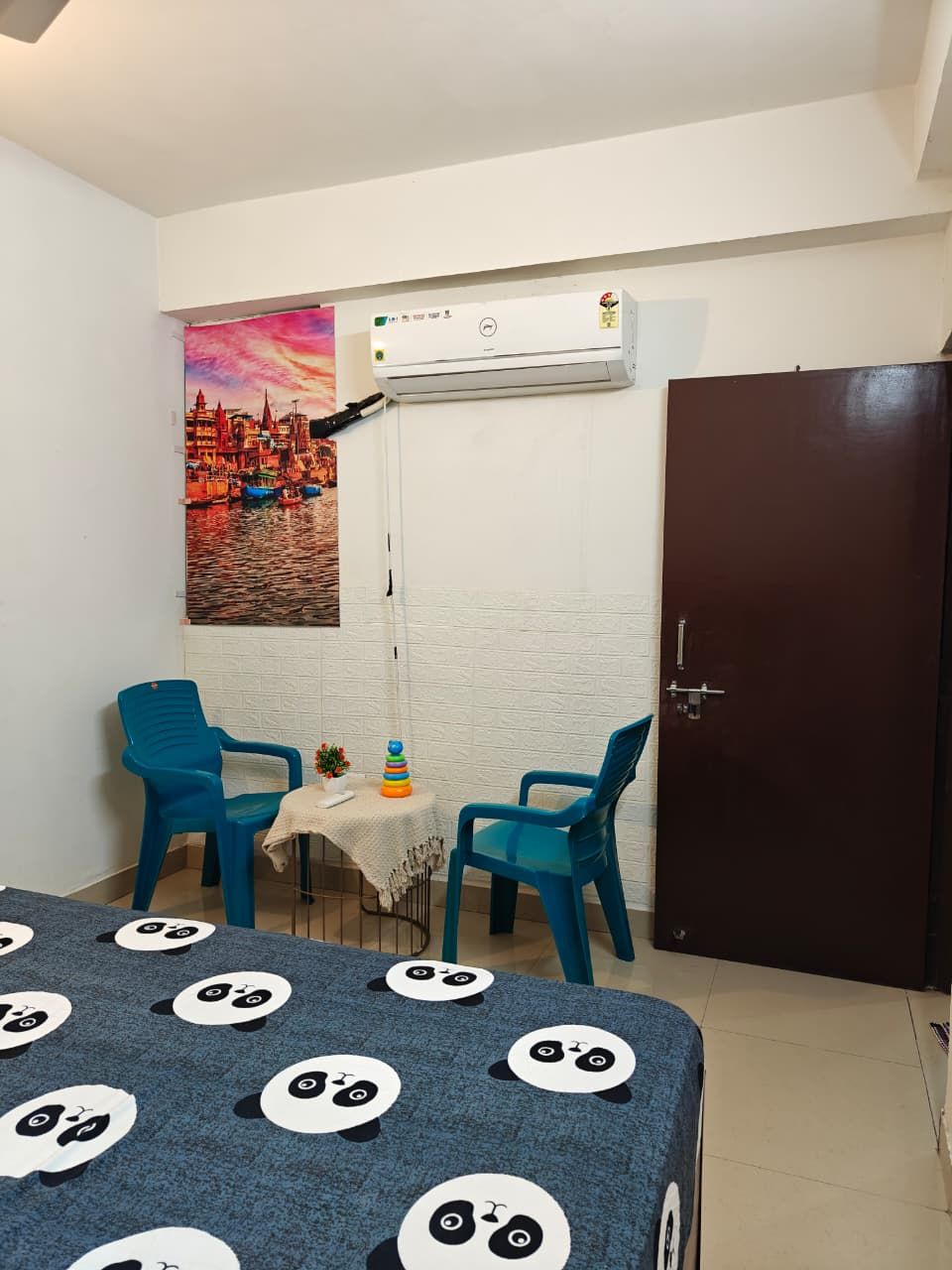 Ein Zimmer mit einem Bett, einem Tisch mit einer Tischdecke mit Panda-Motiv, zwei Stühlen, einer Wand-Klimaanlage und einer Tür.