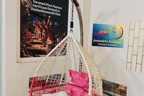 A hanging chair with a netted structure, a poster of a performance, and a sign for Aavimukta Holidays. - Entire 2BHK Spacious Premium Homestay near Kedar Ghat (Varanasi)