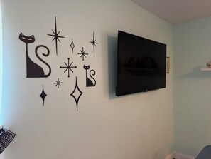 A wall with decorative star and cat silhouettes, a flat-screen TV mounted above a shelf with decorative items, and a partial view of a room with a white baseboard. - No Fees! 1 BR Retro Retreat for 2! Quiet, Cozy town- 15mins from Joplin,Carthage (Alba)