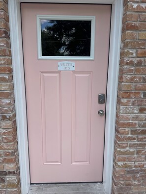 Une porte rose avec une plaque indiquant « Suite 105 » au-dessus, une fenêtre au-dessus de la porte et un interphone sur le côté droit.