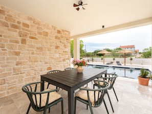 A stone wall, a table with chairs, a flower vase, a ceiling fan, and a pool with umbrellas. - Beautiful Villa Lorna with pool by Interhome (Biograd na Moru)
