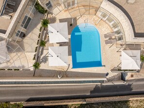 En vy från ovan av en swimmingpool omgiven av solstolar och parasoller, med ett däck av stenplattor och en böjd glasstruktur ovanför.
