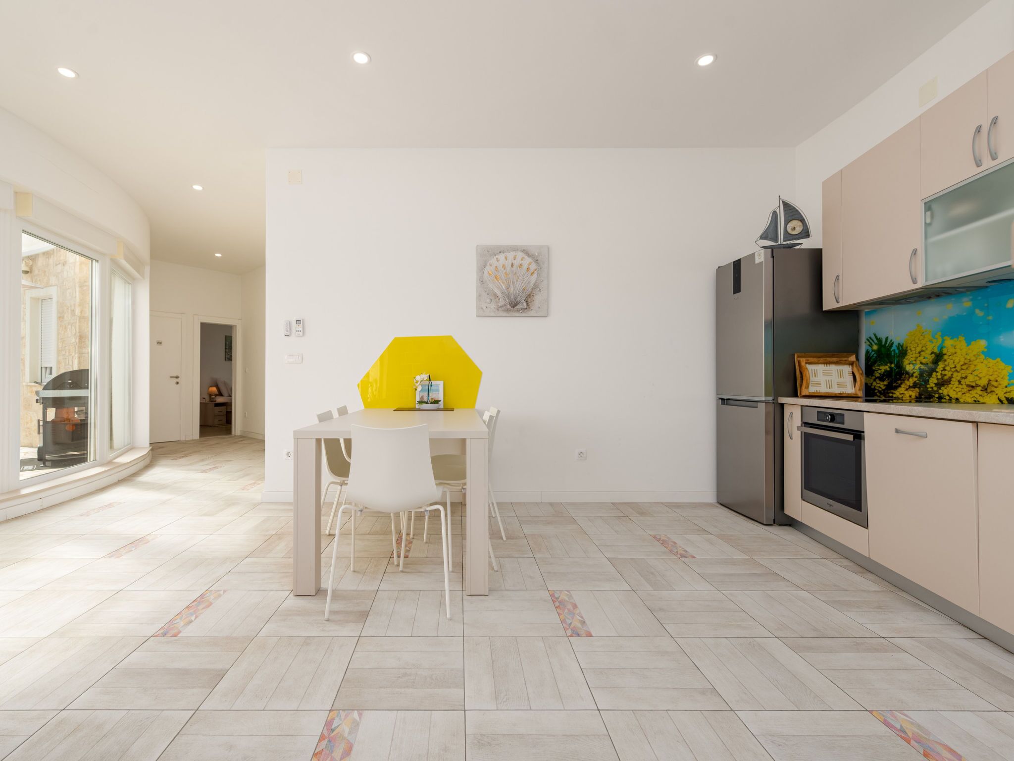 A modern kitchen with a dining area, featuring a large window, a yellow table, and a refrigerator.
