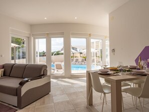 Ein modernes Wohnzimmer mit einer Couch, einem Esstisch für zwei Personen und Blick auf ein Außen-Schwimmbad.