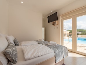Ein Schlafzimmer mit Bett, einem an der Wand befestigten Fernseher und Blick auf einen Swimmingpool durch eine Glastür.