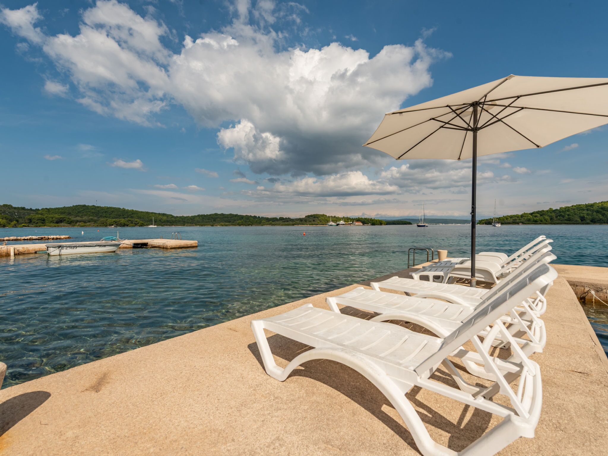 A waterfront area with lounge chairs and an umbrella, a boat docked, and a clear view of the water and distant land.
