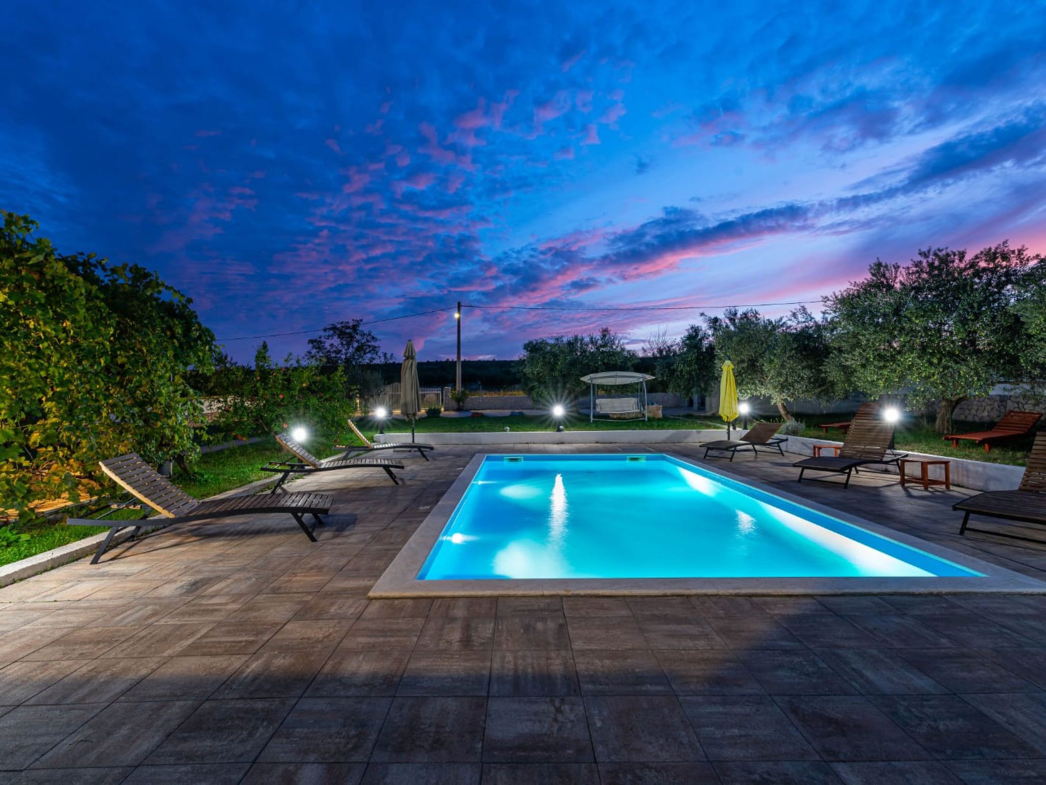 Et rektangulært swimmingpool med fliseterrasse, omgivet af liggestole og parasoller under en skumringsagtig himmel.