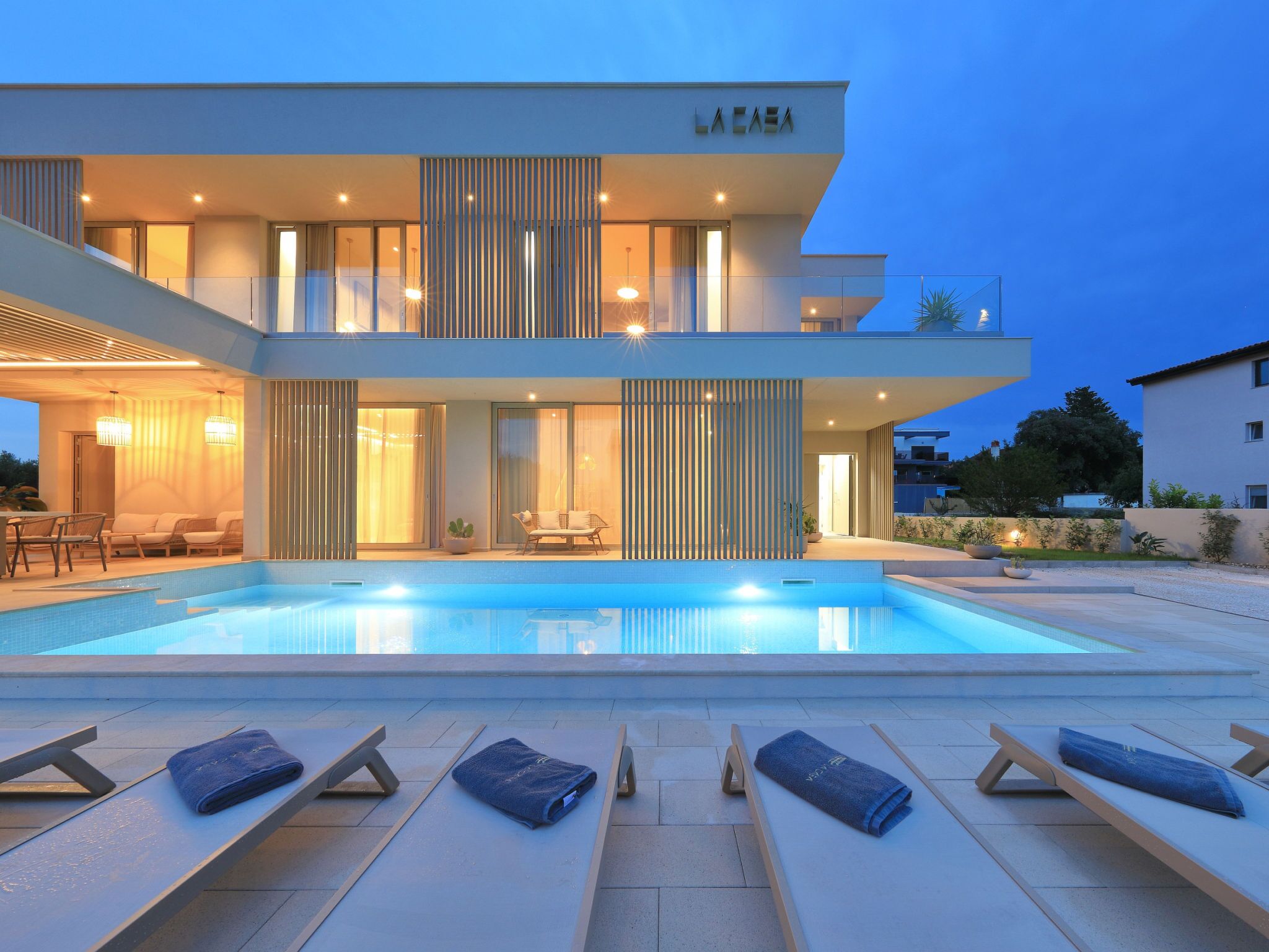 Un bâtiment moderne à deux étages, doté d’une piscine, de chaises longues et d’un intérieur bien éclairé.