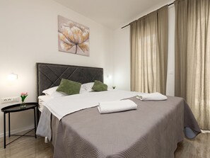 Ein Hotelzimmer mit Bett, Nachttisch, Wandlampe und großem Fenster mit Vorhängen.
