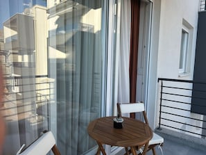 A balcony with a wooden chair and table, a window with curtains, and a view of buildings. - Apartment  by Interhome (Zadar)