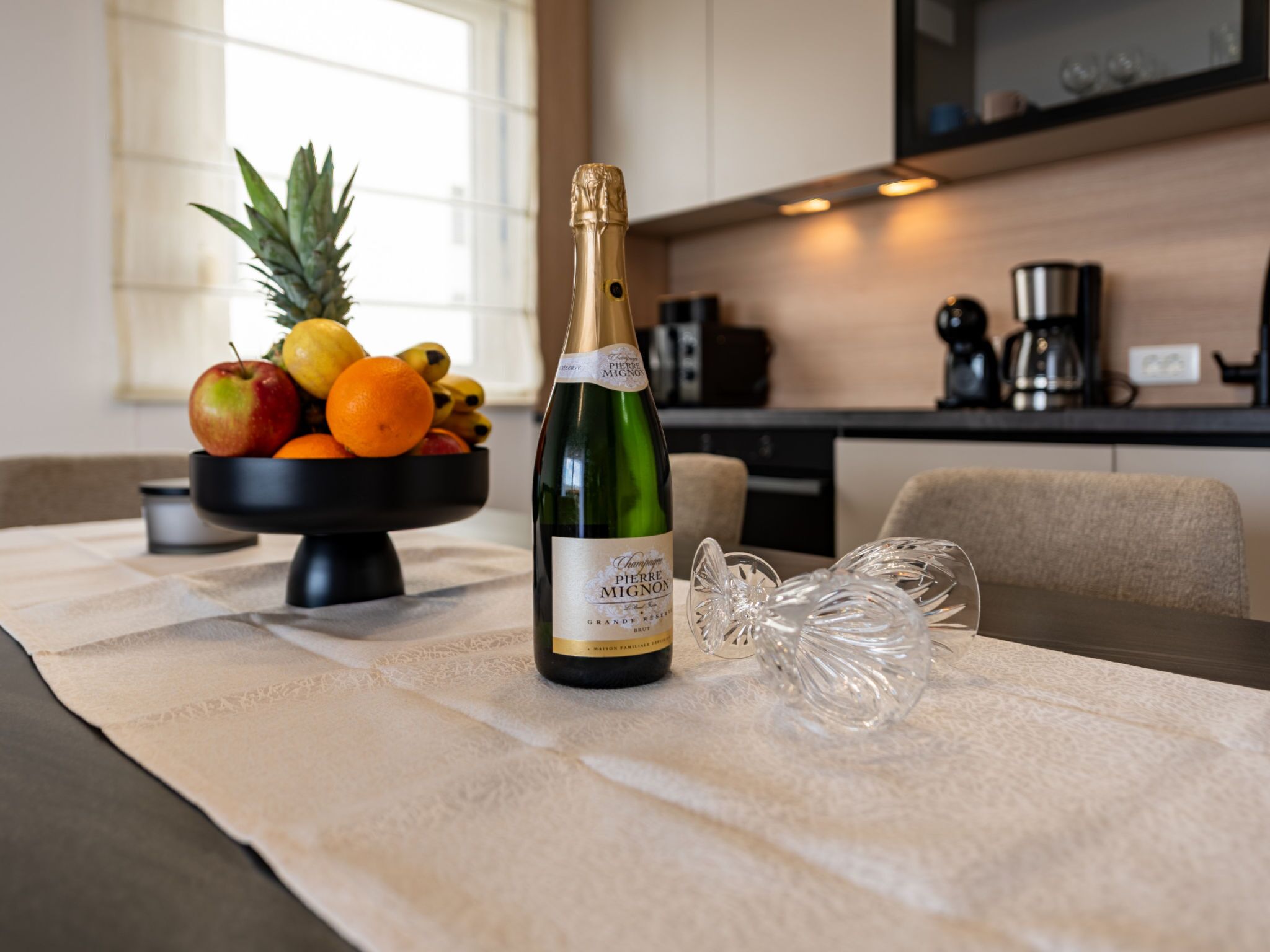 A bottle of champagne, a fruit bowl, and a glass on a table in a modern kitchen.