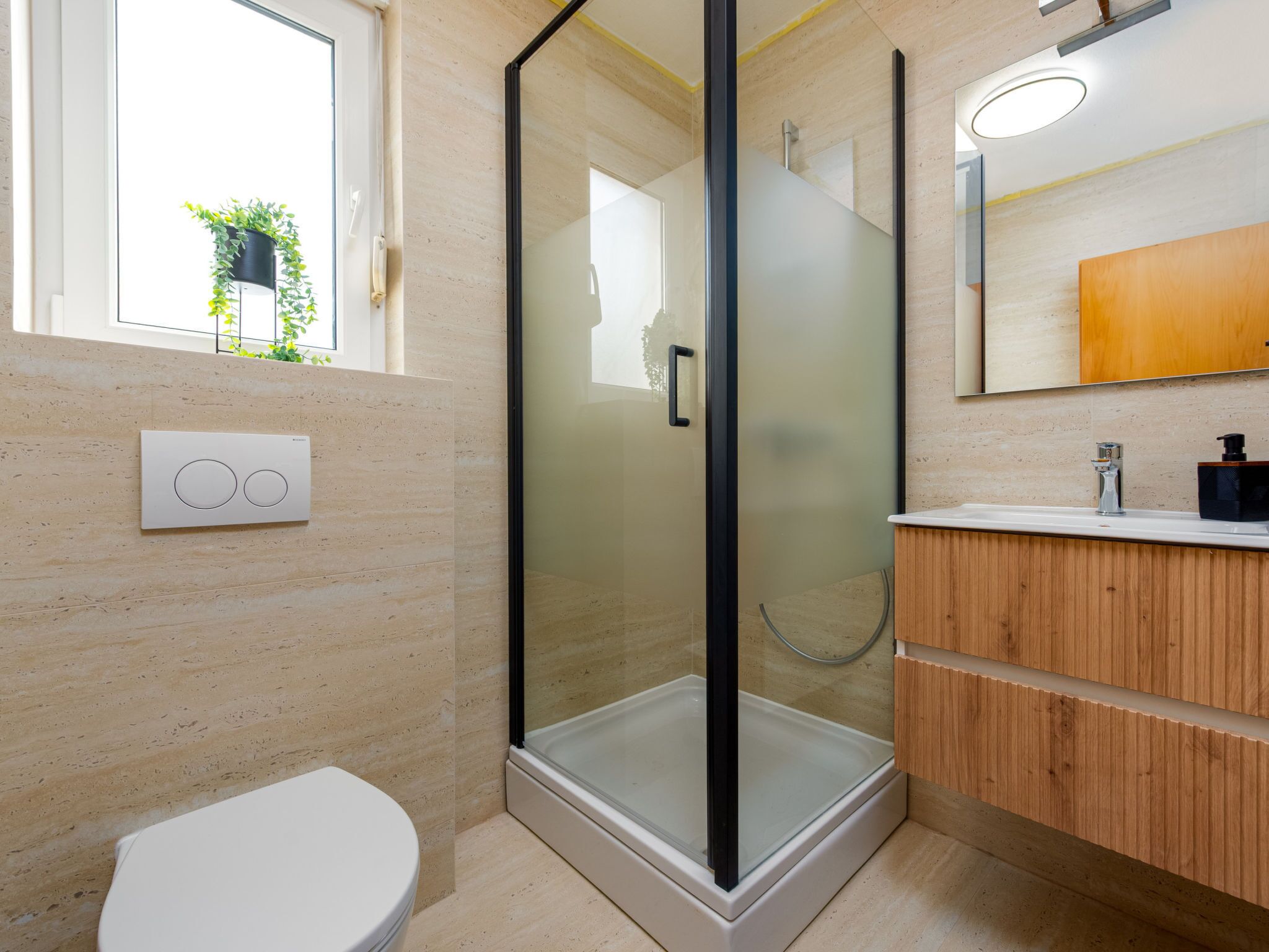 A modern bathroom with a glass shower enclosure, a toilet, a vanity with a mirror, and a window with a plant.