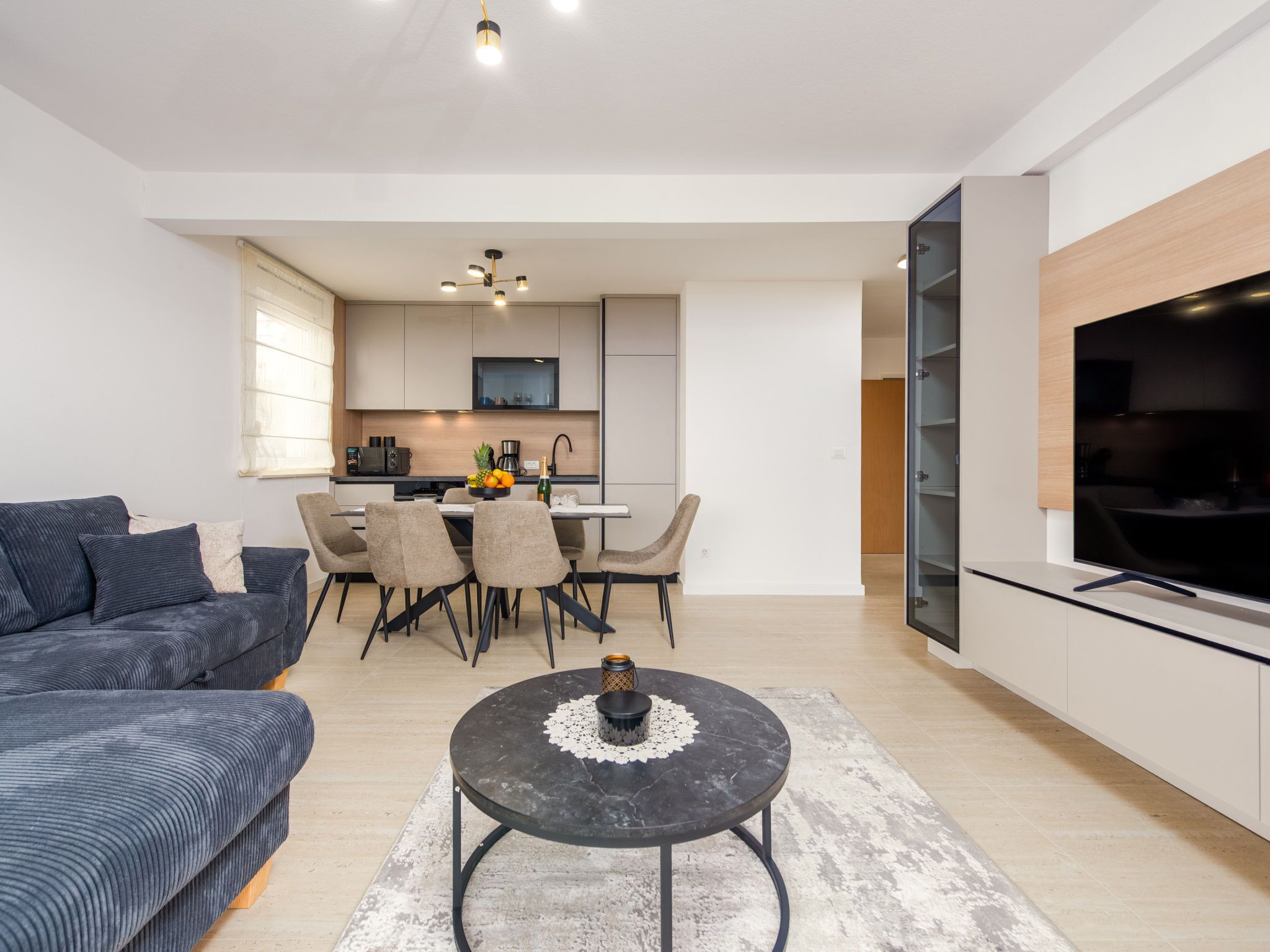 A modern living room with a sofa, a dining area with chairs and a table, a kitchen with cabinets and appliances, and a TV on a stand.