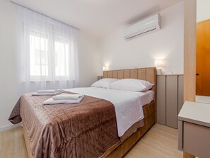 Ein Hotelzimmer mit Bett, Nachttisch, Klimaanlage und einem Fenster mit durchsichtigen Vorhängen.