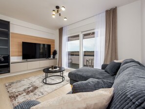 Ein modernes Wohnzimmer mit großem Flachbildfernseher, einem Ecksofa, einem Couchtisch und Blick auf einen Balkon.