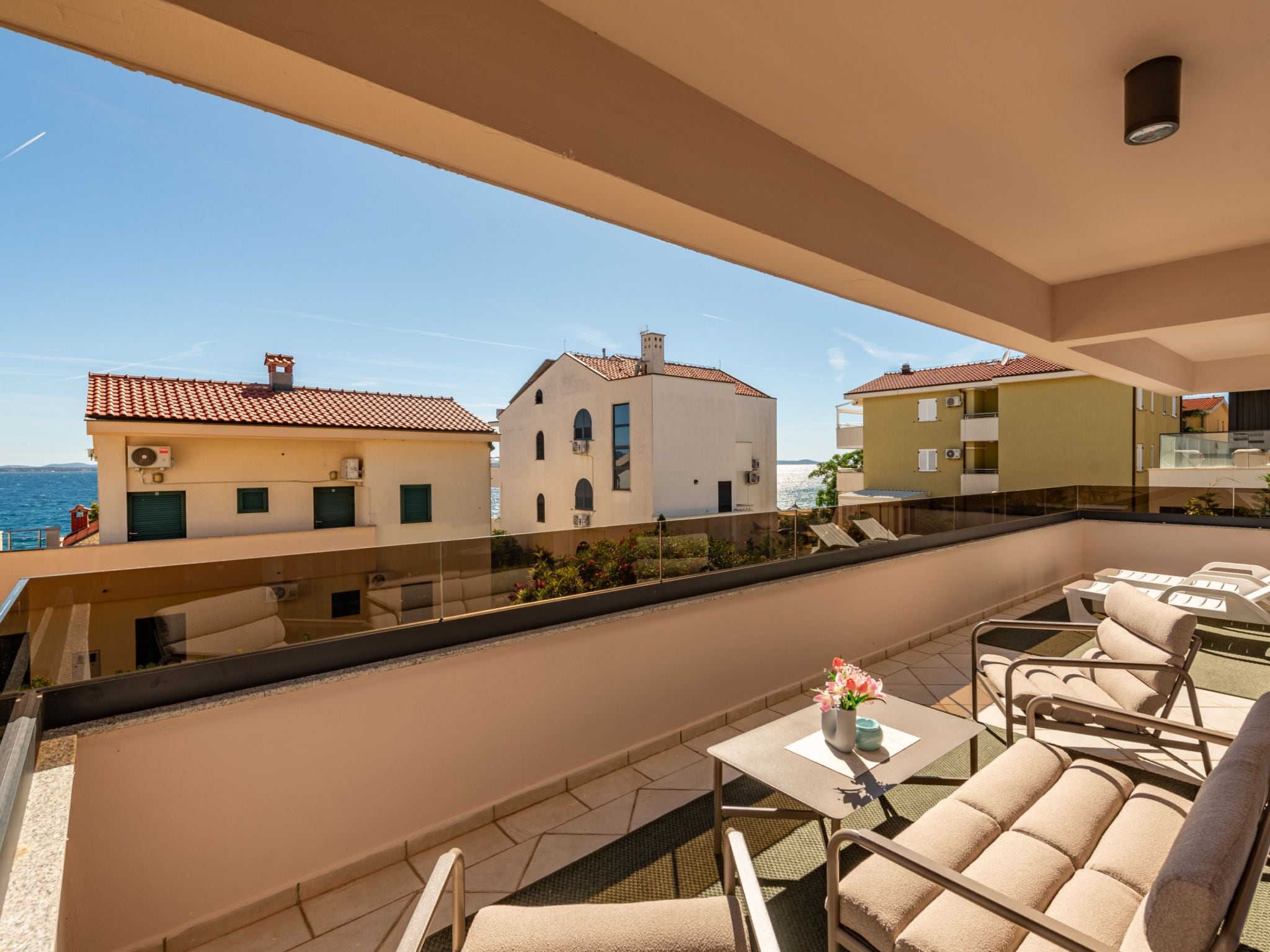 A balcony with outdoor seating, a view of the sea, and buildings with red-tiled roofs.