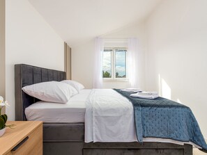 A bedroom with a bed, white bedding, a blue accent bedspread, a window with curtains, and a view of the sky. - Seaview apartment Casa Almaris A3 by Interhome (Zadar/Kožino)