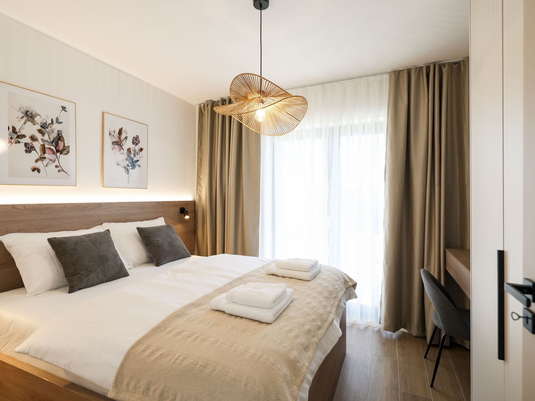A hotel room with a large bed, two framed floral artworks, a hanging light fixture, and a view of the outdoors through sheer curtains.