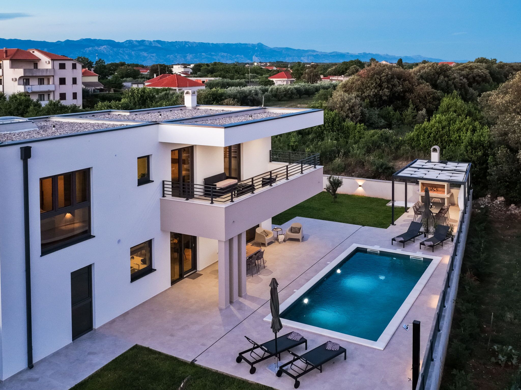 A modern two-story house with a swimming pool, outdoor seating, and a view of the mountains.