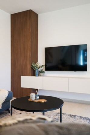 Ein modernes Wohnzimmer mit einem runden, schwarzen Couchtisch, einem flachbildfernseher an einer weißen Wand und einer Wand in Holzoptik.