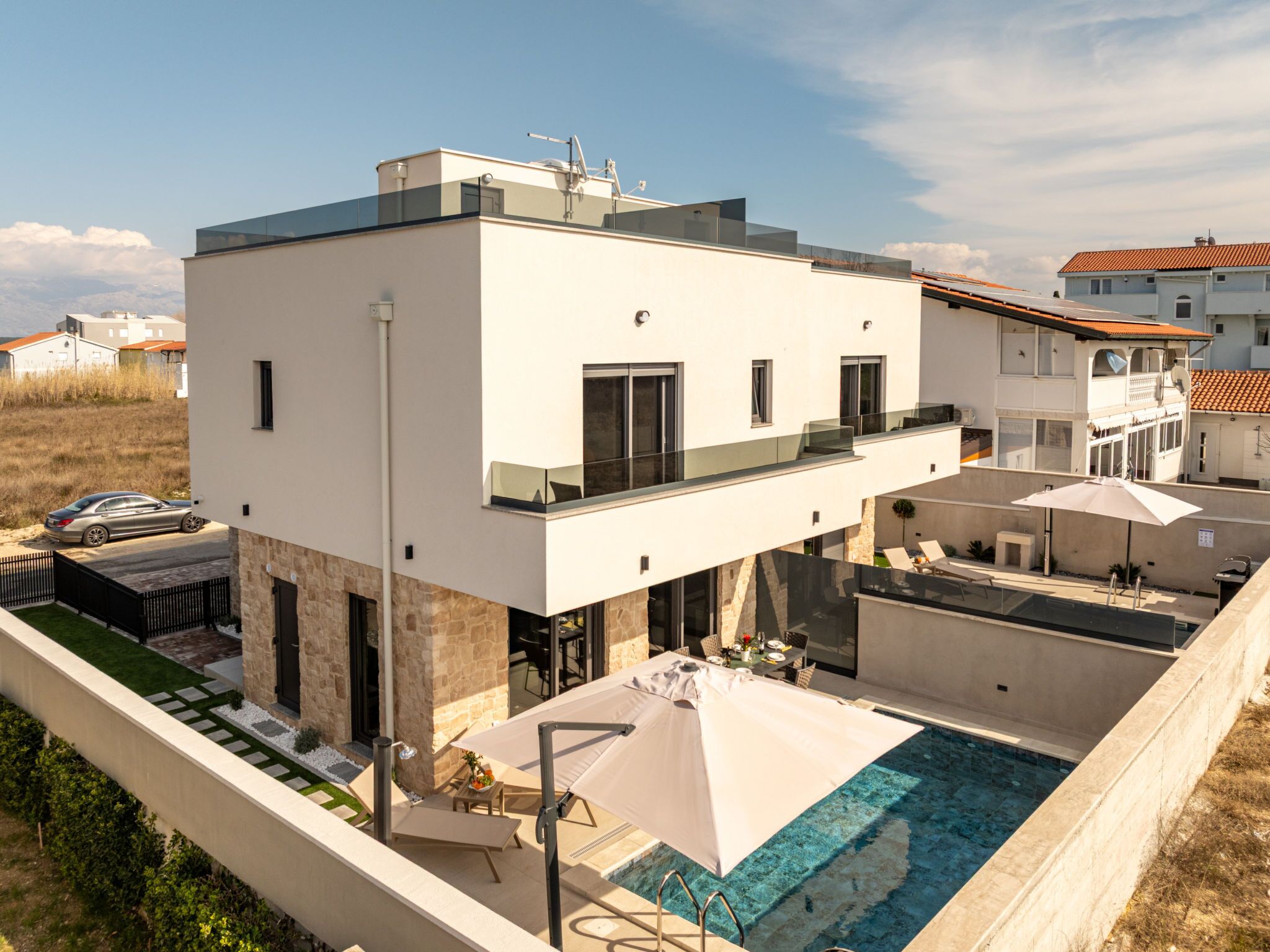 A modern two-story house with a rooftop pool, outdoor seating, and a car parked nearby.