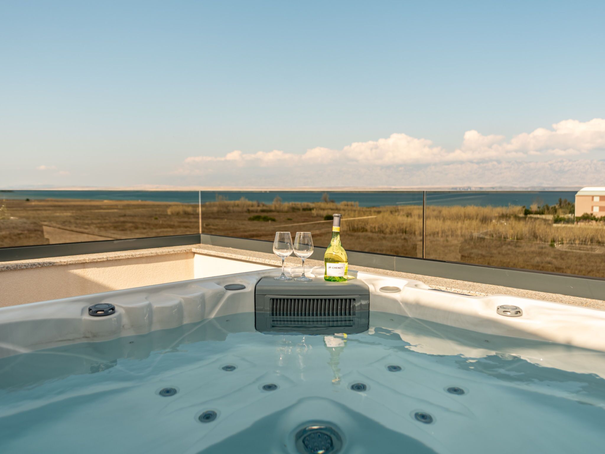 A hot tub with a bottle of wine and glasses on a balcony overlooking a beach.