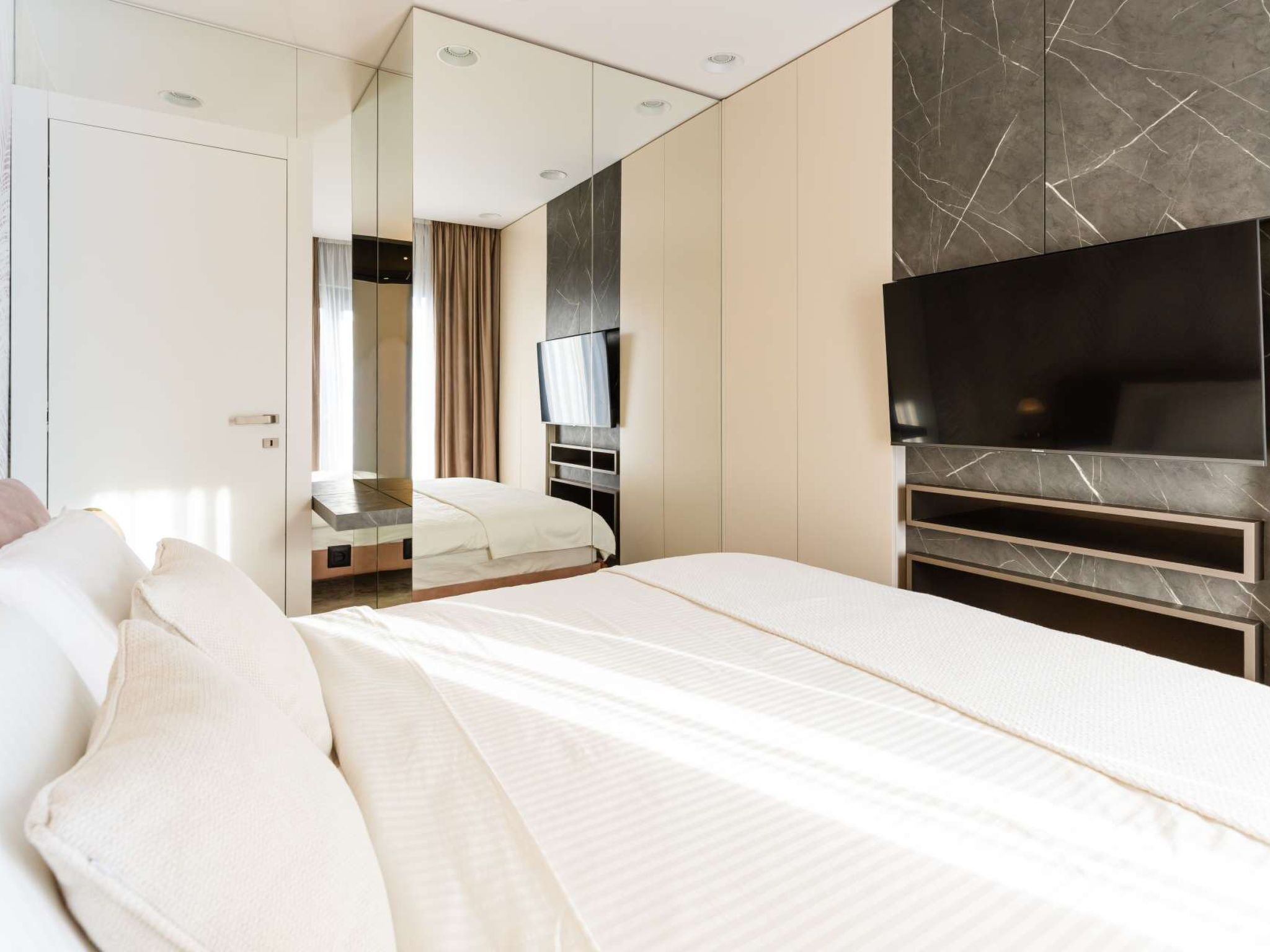 A modern hotel room with a large bed, a flat-screen TV mounted on a marble wall, and a wardrobe with a mirror.