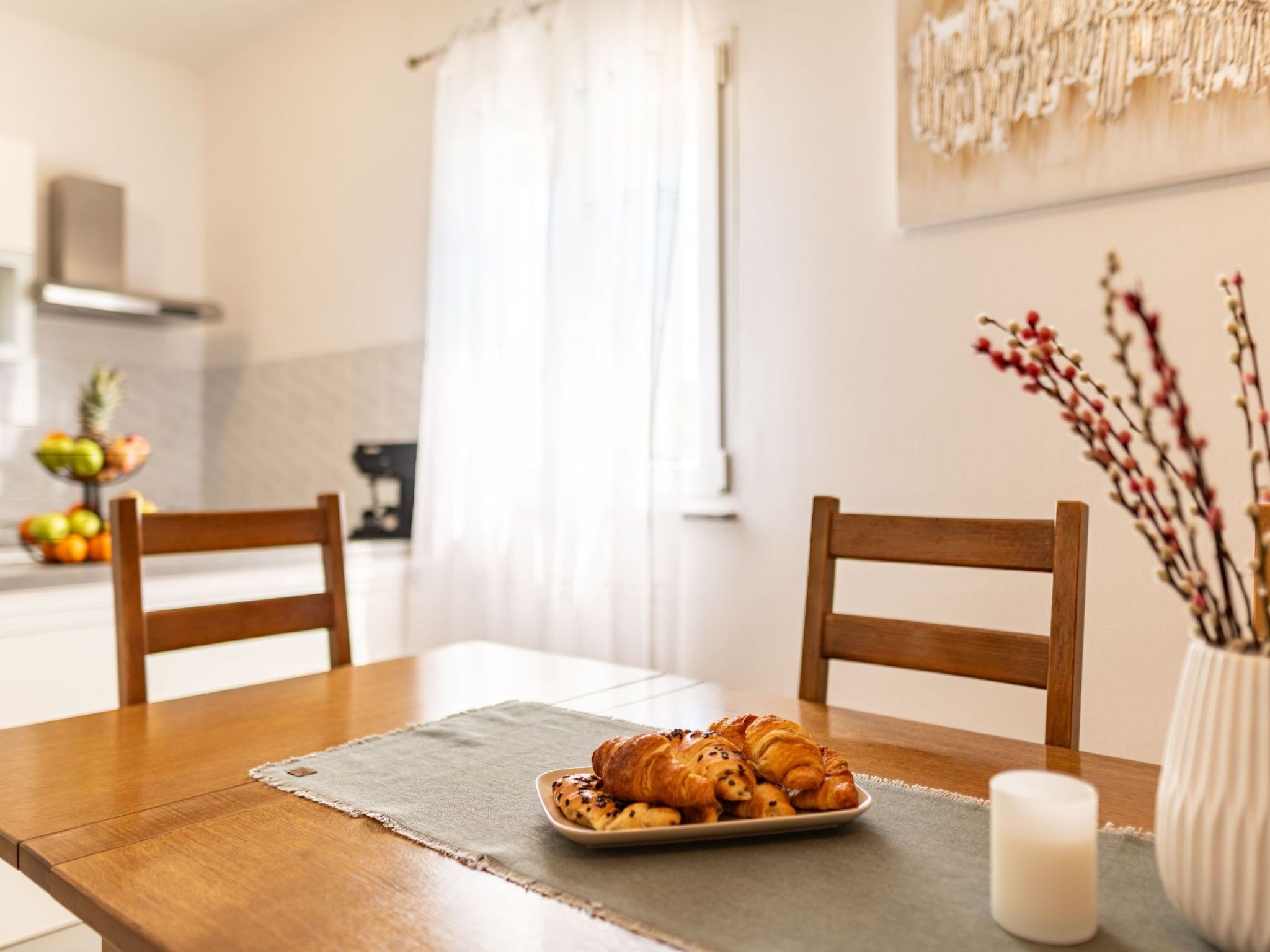 A dining table with a plate of pastries and a candle, two wooden chairs, a vase with branches, and a kitchen in the background.