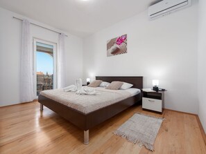 Ein modernes Schlafzimmer mit einem großen Bett, Nachttischen, einem Schreibtisch und Blick auf die Stadt durch ein Fenster, sowie einer Klimaanlage.