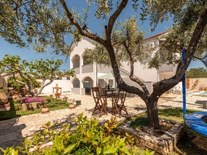 Ein Innenhof mit Tisch und Stühlen, umgeben von Olivenbäumen und einem Gebäude mit Bögen.
