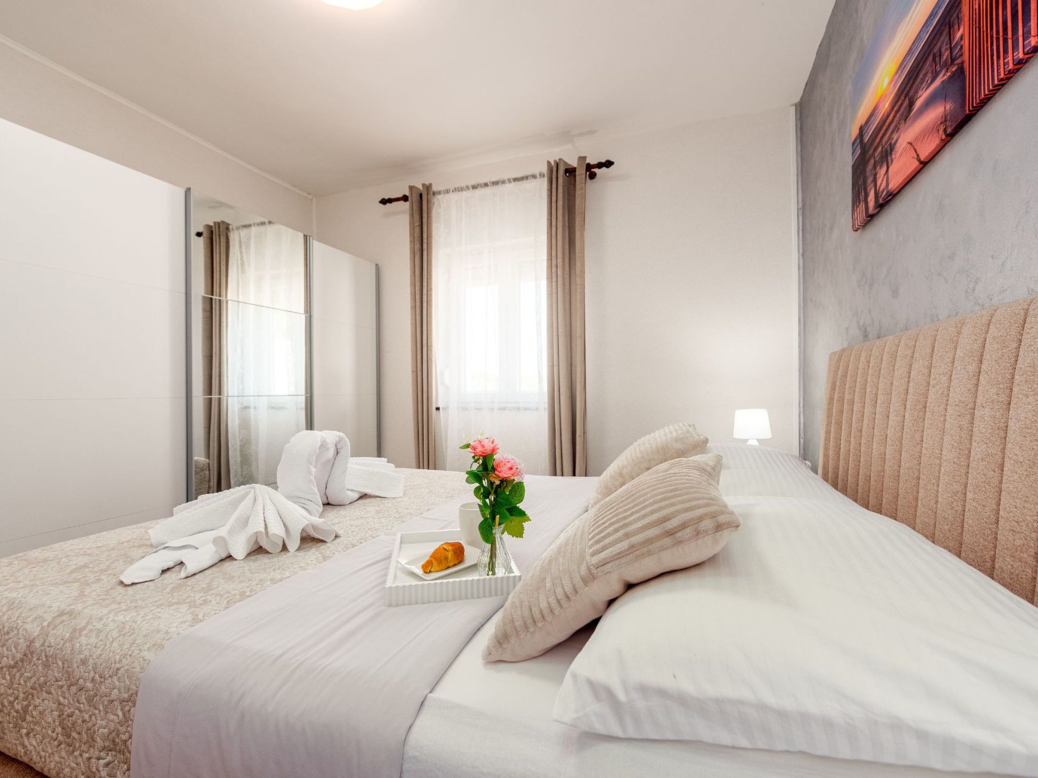 A hotel room with a bed, a bedside table with a plate of food and flowers, and a large window with curtains.