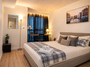 A hotel room with a bed, bedside tables, a desk, a chair, a potted plant, and a painting of a cityscape. - The Imperial Waterfront Escape (Toronto)