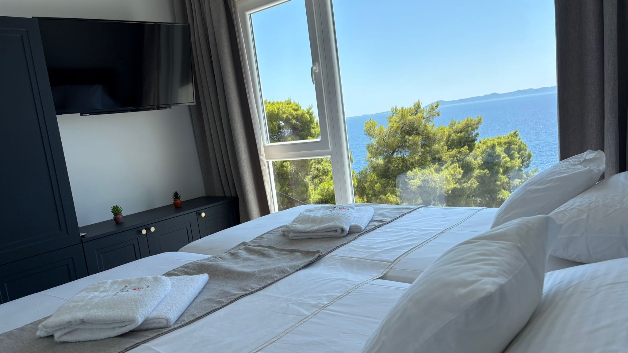 A bedroom with a bed, a TV, a window with a view of the sea, and a nightstand with a plant.