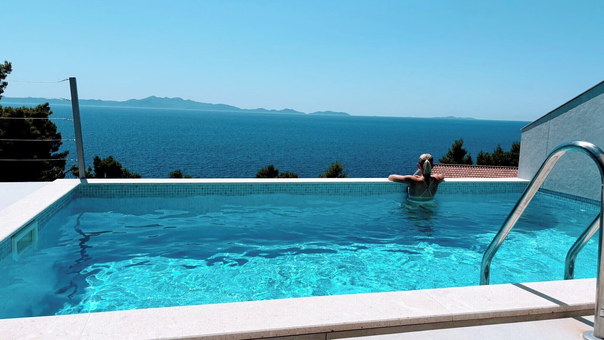 A swimming pool with clear blue water, a metal ladder, and a view of the sea and distant mountains.