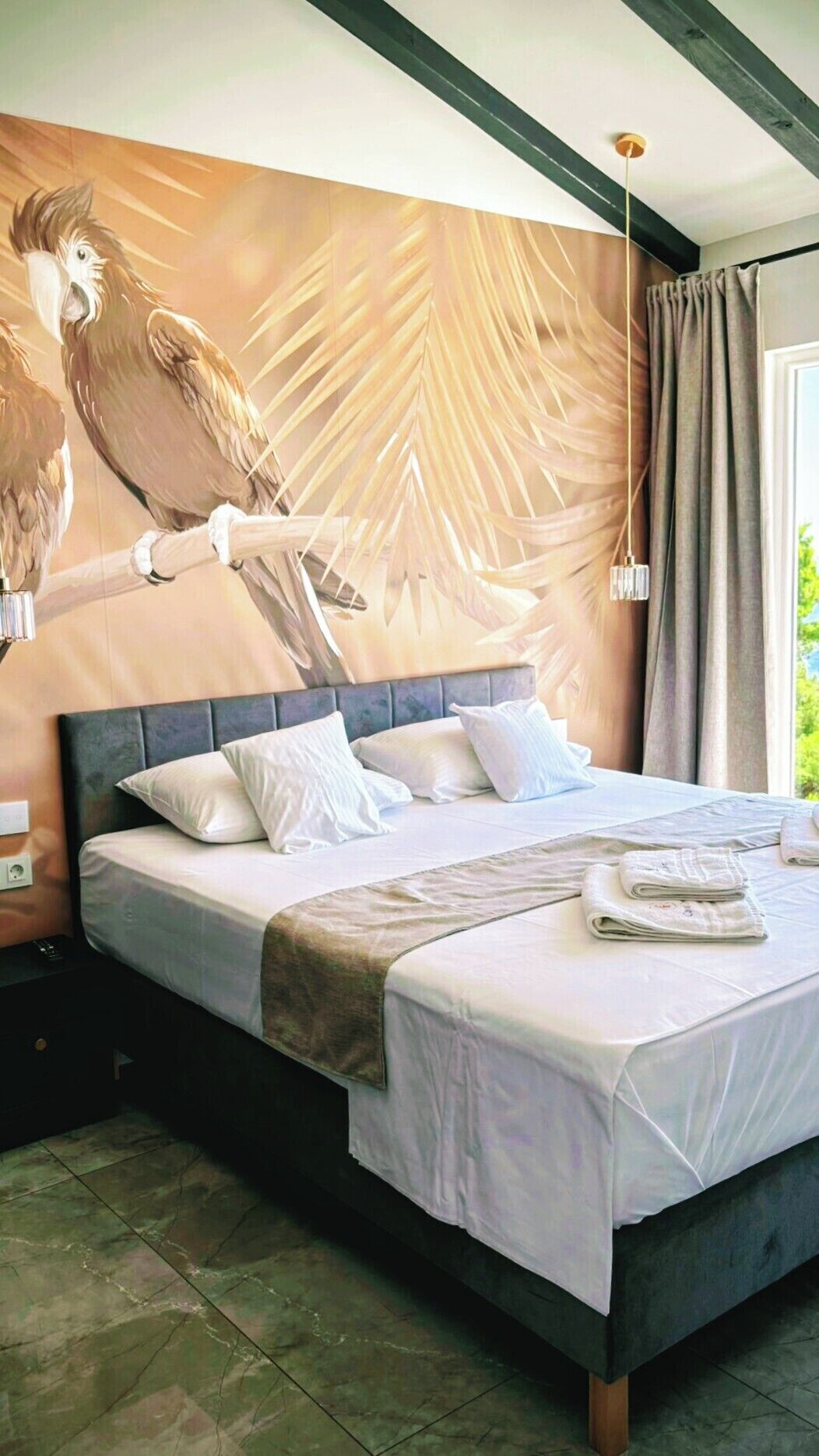 Un dormitorio con una cama grande, cabecera, mesita de noche, un mural en la pared y una ventana con cortinas.