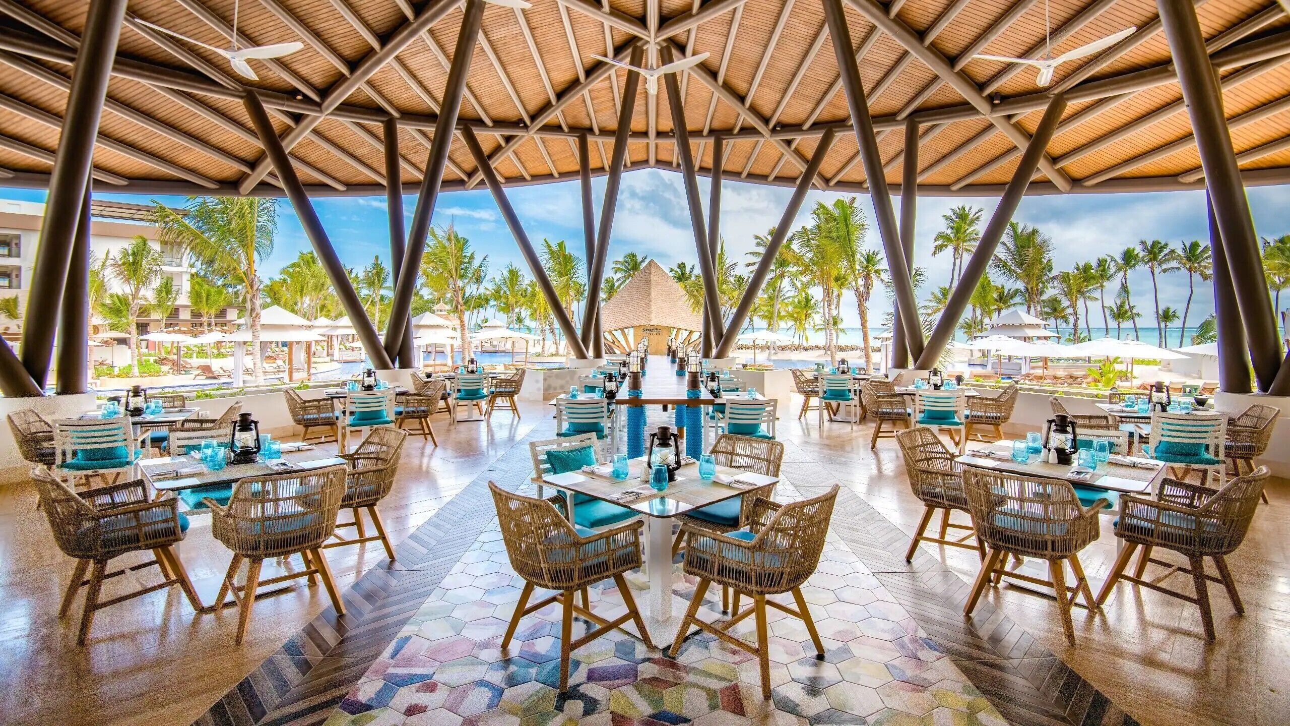 Un vaste espace extérieur aménagé pour les repas, avec plusieurs tables et chaises, un toit de chaume et une vue sur des palmiers et une plage.