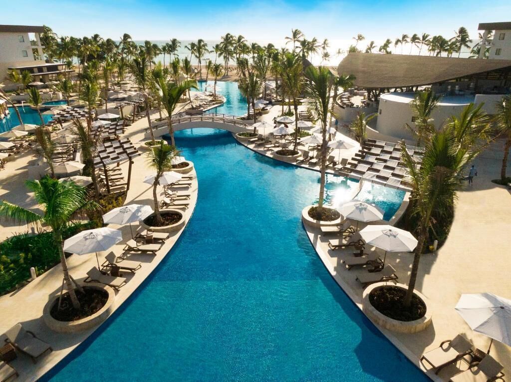Un espace piscine dans un complexe hôtelier, comprenant plusieurs piscines, des chaises longues et des palmiers.