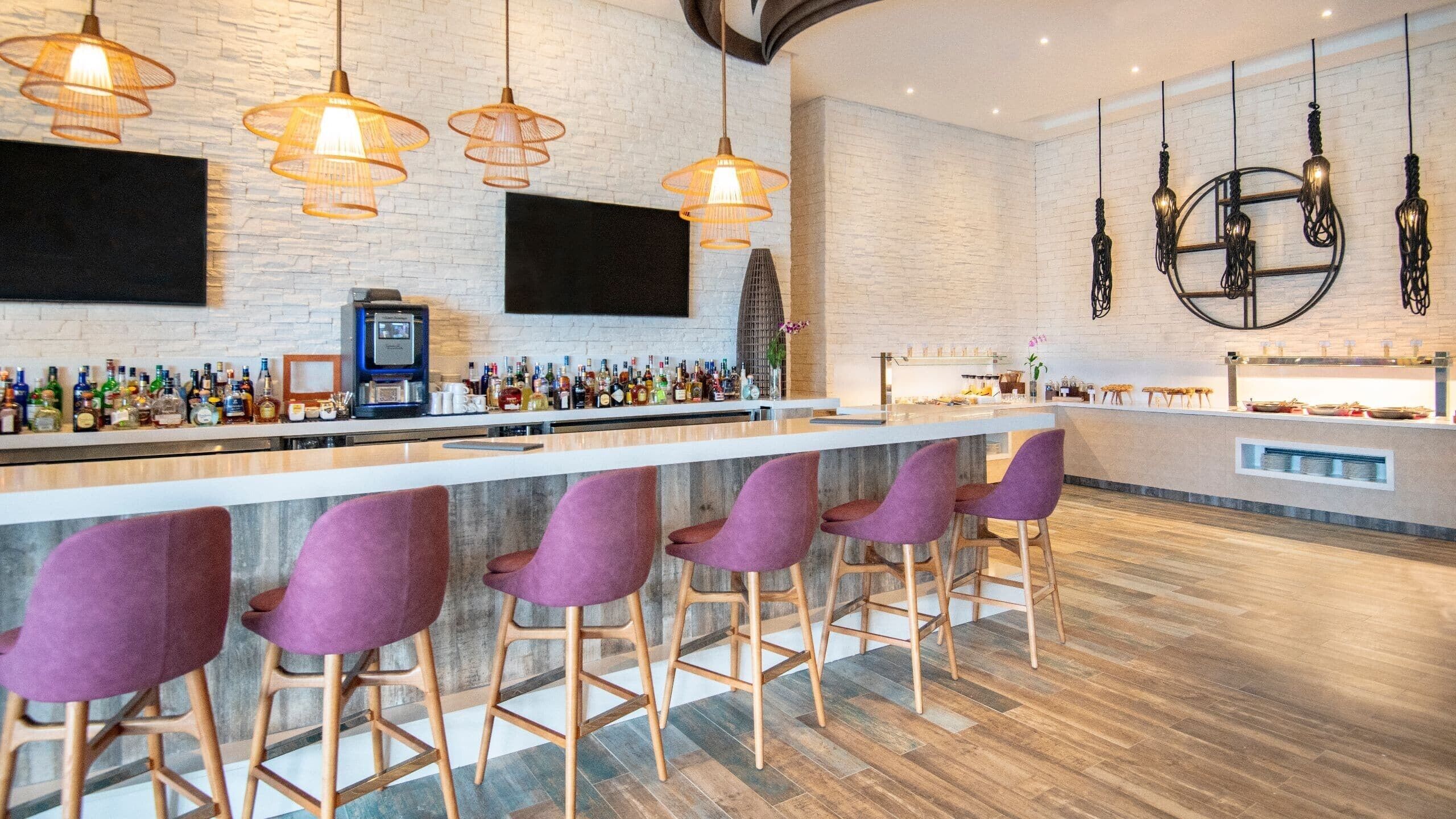 A bar area with a long counter, multiple liquor bottles, a coffee machine, and purple bar stools.