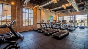 Un gimnasio con varias cintas de correr dispuestas en filas, ventanas amplias y una pared de ladrillo.