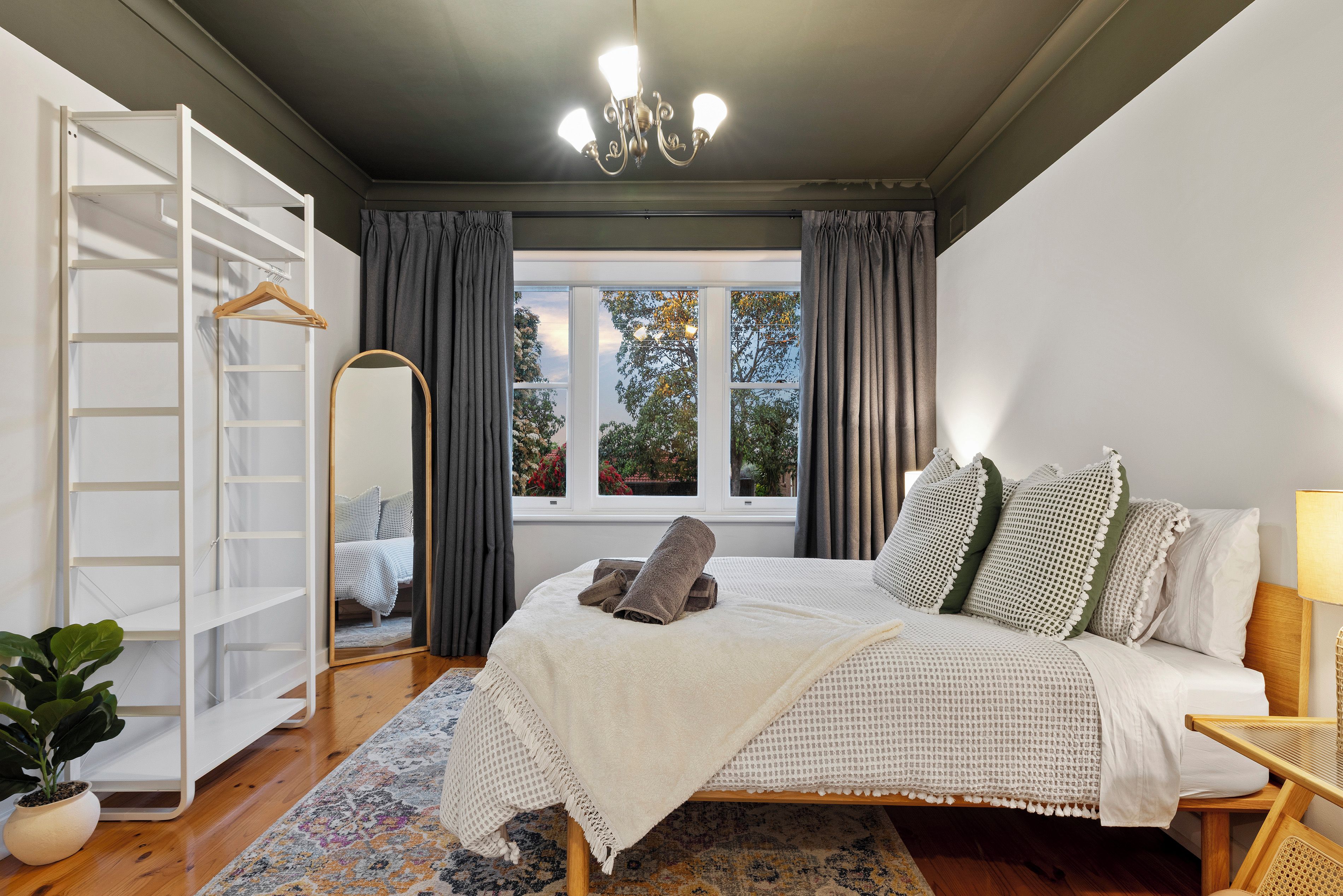 A bedroom with a bed, a bedside table, a lamp, a wardrobe, a mirror, and a window with curtains.