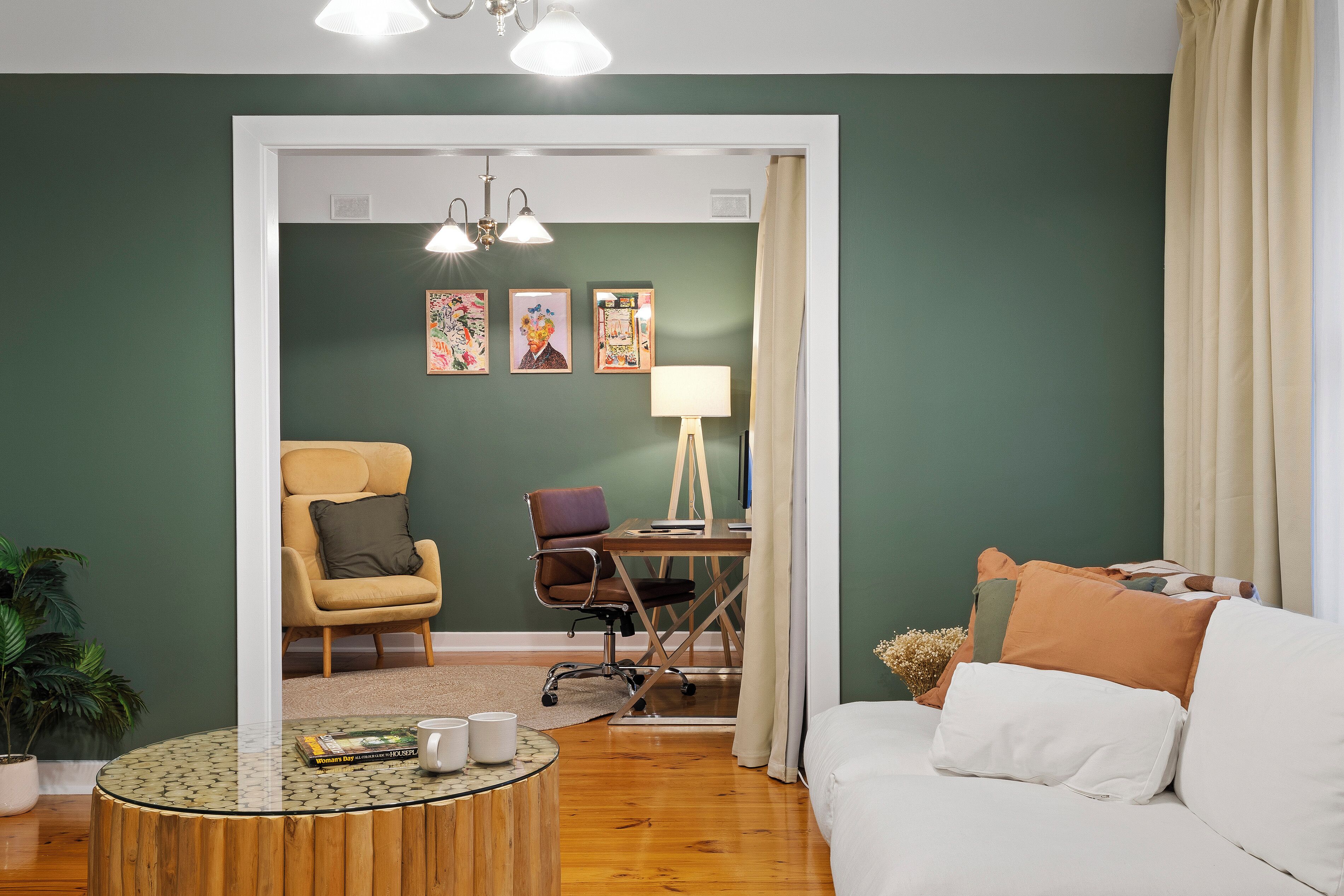 Een moderne woonkamer met een bank, fauteuils, een salontafel en een bureau met een lamp. Er hangen ingelijste kunstwerken aan de muur en staat een potplant op de vloer.