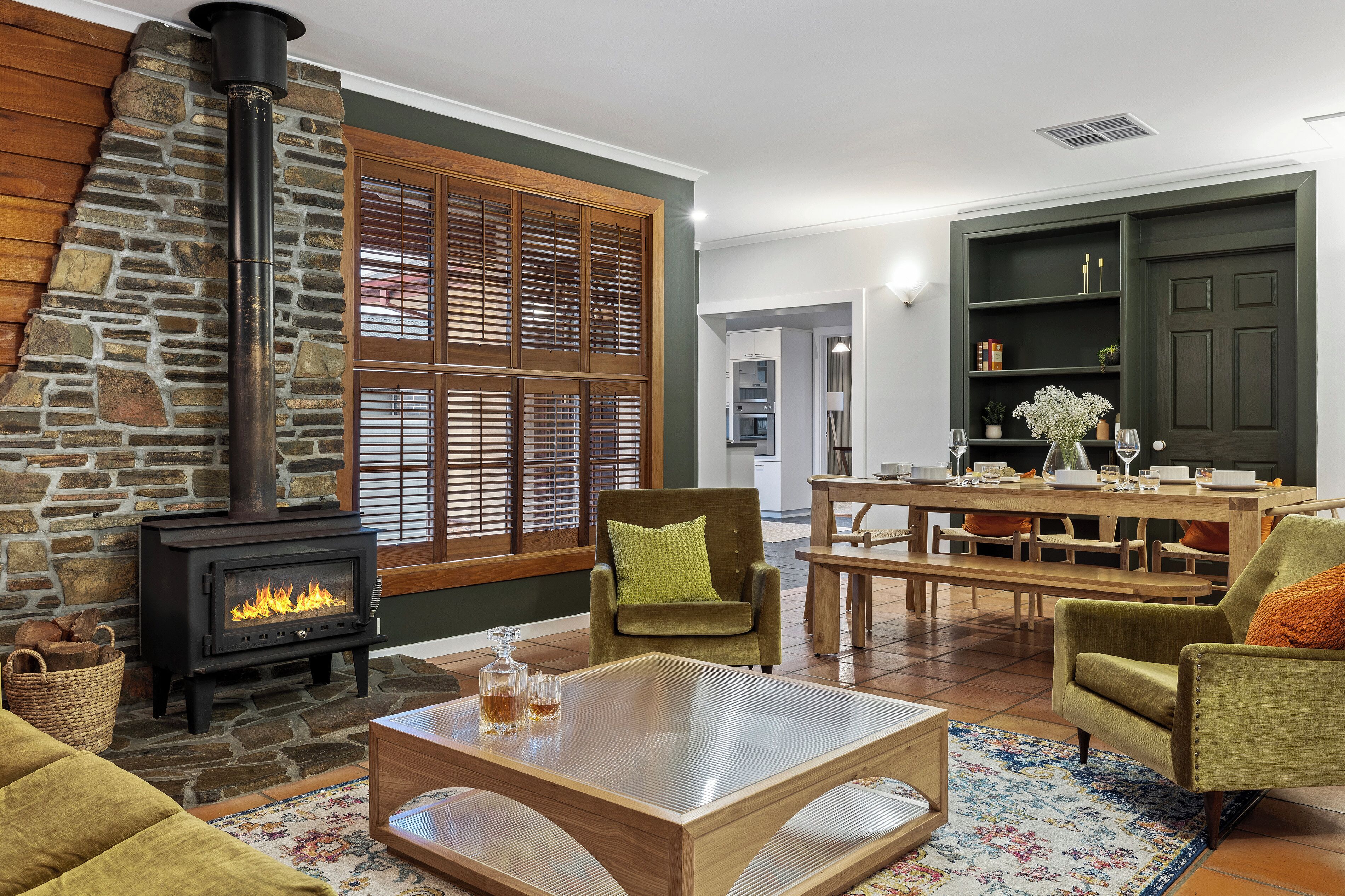 A cozy living room with a stone fireplace, wooden blinds, and a dining area with a table and chairs.