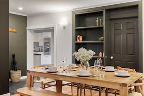 A dining area with a wooden table set for four, chairs, a vase with flowers, and a kitchen in the background.