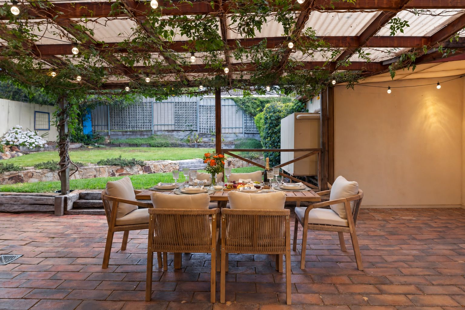 An outdoor patio with a table set for four, surrounded by wicker chairs, under a pergola with string lights.