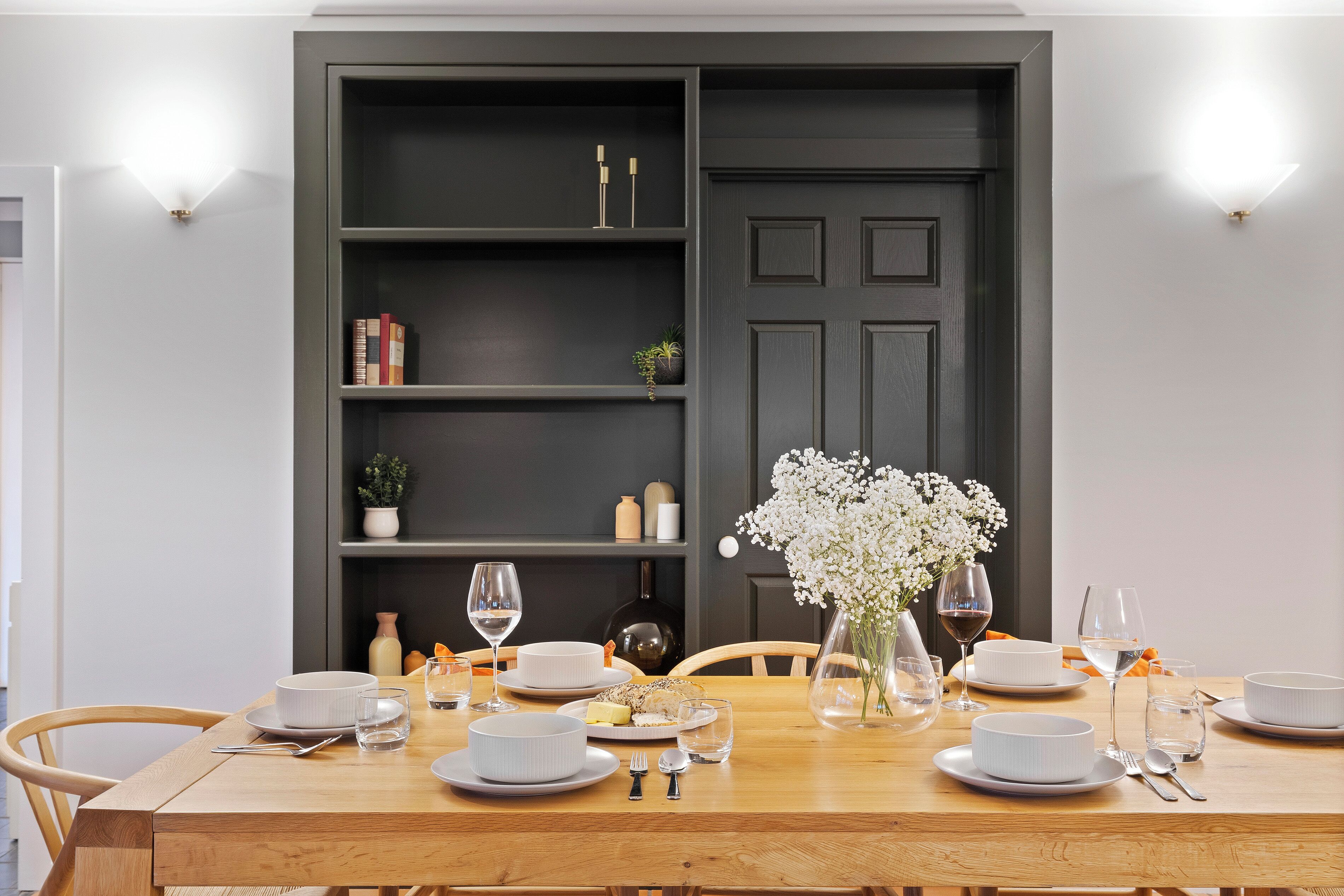 Un coin repas avec une table en bois dressée pour quatre personnes, entourée de chaises. On y trouve une bibliothèque remplie de livres et d’objets décoratifs, ainsi qu’une porte sombre au fond.