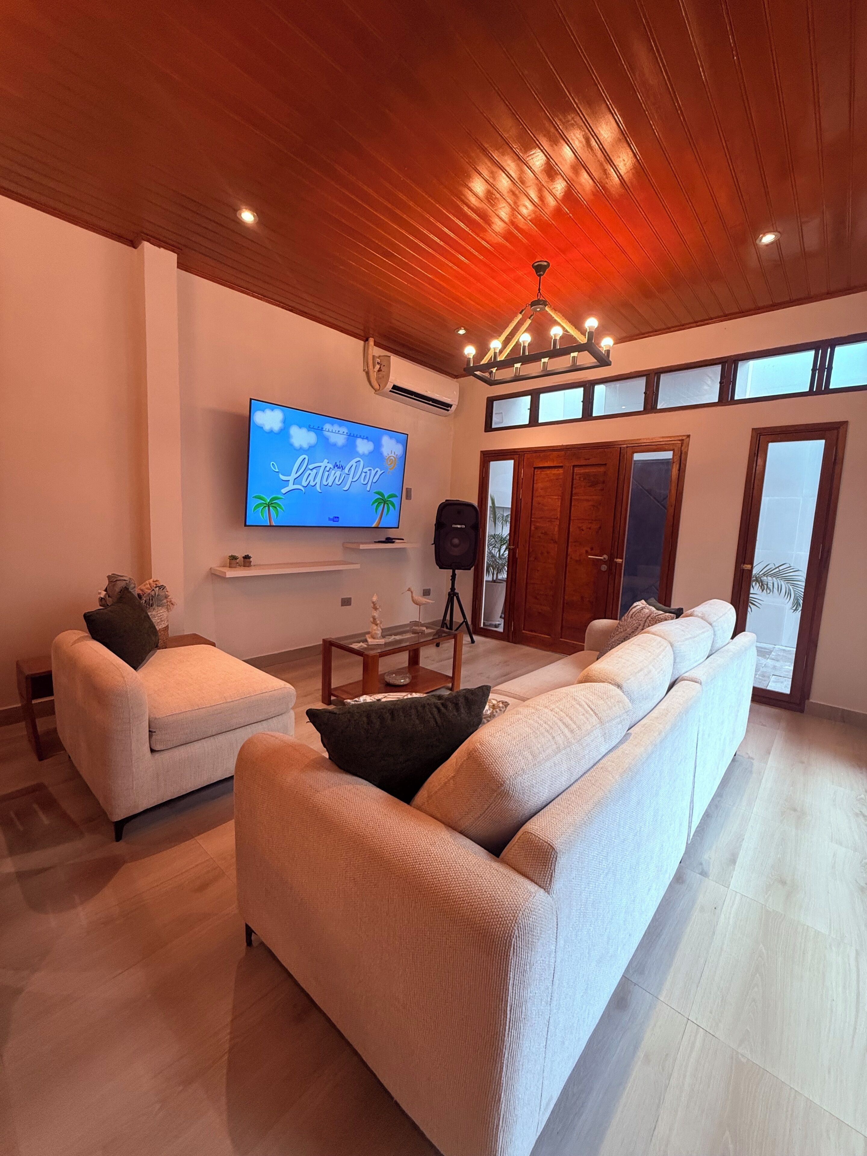 A living room with a large flat-screen TV, a wooden ceiling, and a modern chandelier.